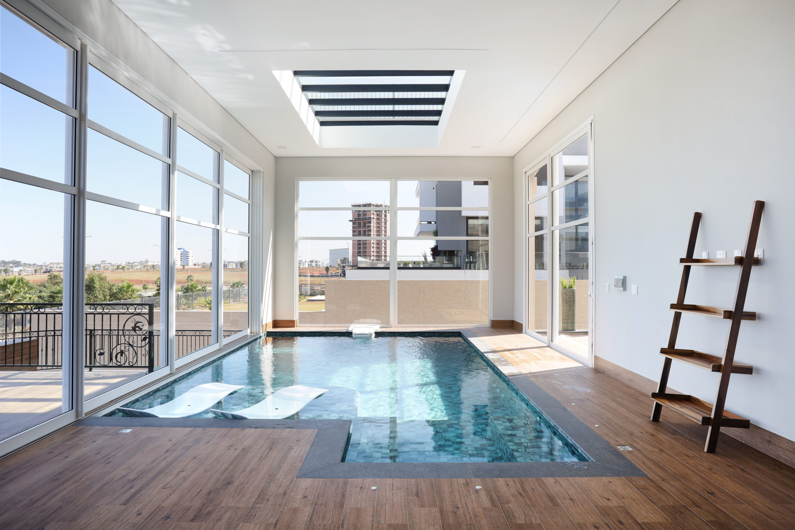 Uma piscina localizada no ambiente interno da casa, o espaço possui janelas grandes e uma vista para a piscina