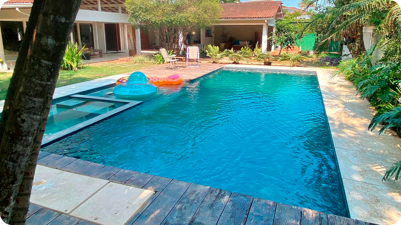 Piscina de alvenaria grande no quintal de uma casa cercada por um jardim. A imagem se relaciona ao artigo sobre os diversos tipos de piscinas